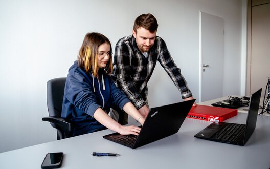 Ausbilder Lucas gemeinsam mit Alex an ihrem zukünftigen Arbeitsplatz als Auszubildende zur Fachinformatikerin für Systemintegration. 